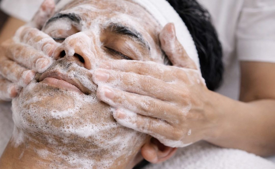 Close-up spa treatment showing a client receiving a cleansing facial Winnipeg service, with an esthetician applying foamy cleanser from behind the head, focusing on professional technique and relaxation.