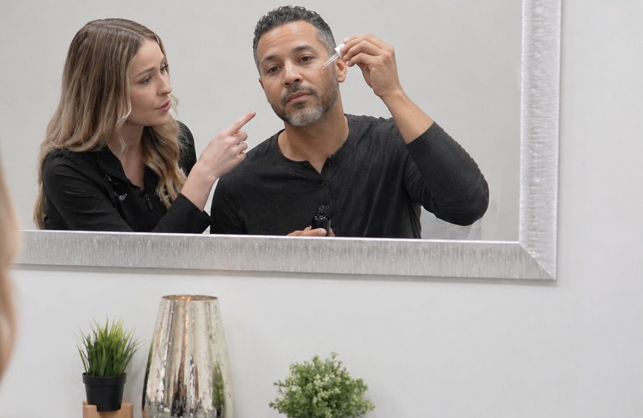 A Winnipeg skin specialist stands beside a client in a modern consultation space, both facing a large wall-mounted mirror. The client’s face is visible in the mirror reflection as they apply a serum to their cheek using a glass dropper, holding the bottle in the other hand. The specialist, positioned slightly behind and to the side, points toward the application area, guiding technique and placement. The room features a sleek cabinet beneath the mirror, styled with small potted plants and a metallic decorative vase. Soft, even lighting and neutral tones create a calm, professional environment focused on personalized skincare instruction.