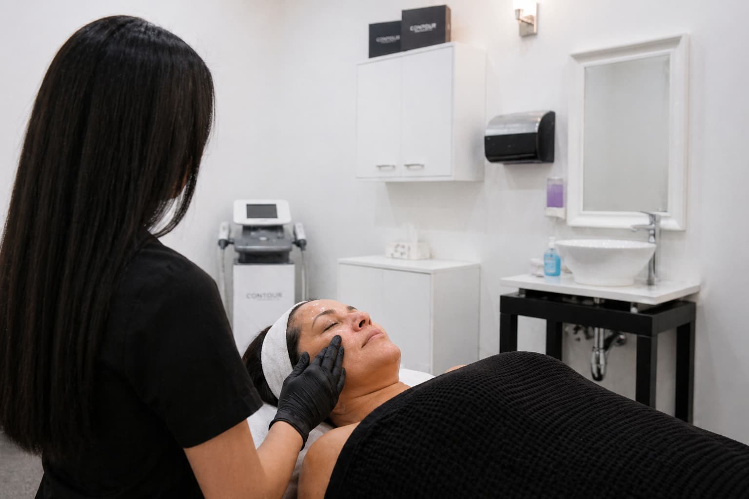 A Winnipeg skin specialist performs a facial treatment on a client in a bright, modern spa room. The practitioner is seen from behind, standing beside a treatment bed and wearing black attire and gloves while gently applying product to the client’s cheek. The client lies comfortably on the bed with a headband holding their hair back and a dark textured blanket covering their body, eyes closed in a relaxed state. The clean, clinical setting includes white walls, soft overhead lighting, a sink with a mirror, mounted dispensers, cabinetry, and professional skincare equipment in the background, creating a calm and organized environment focused on advanced skin care treatments.