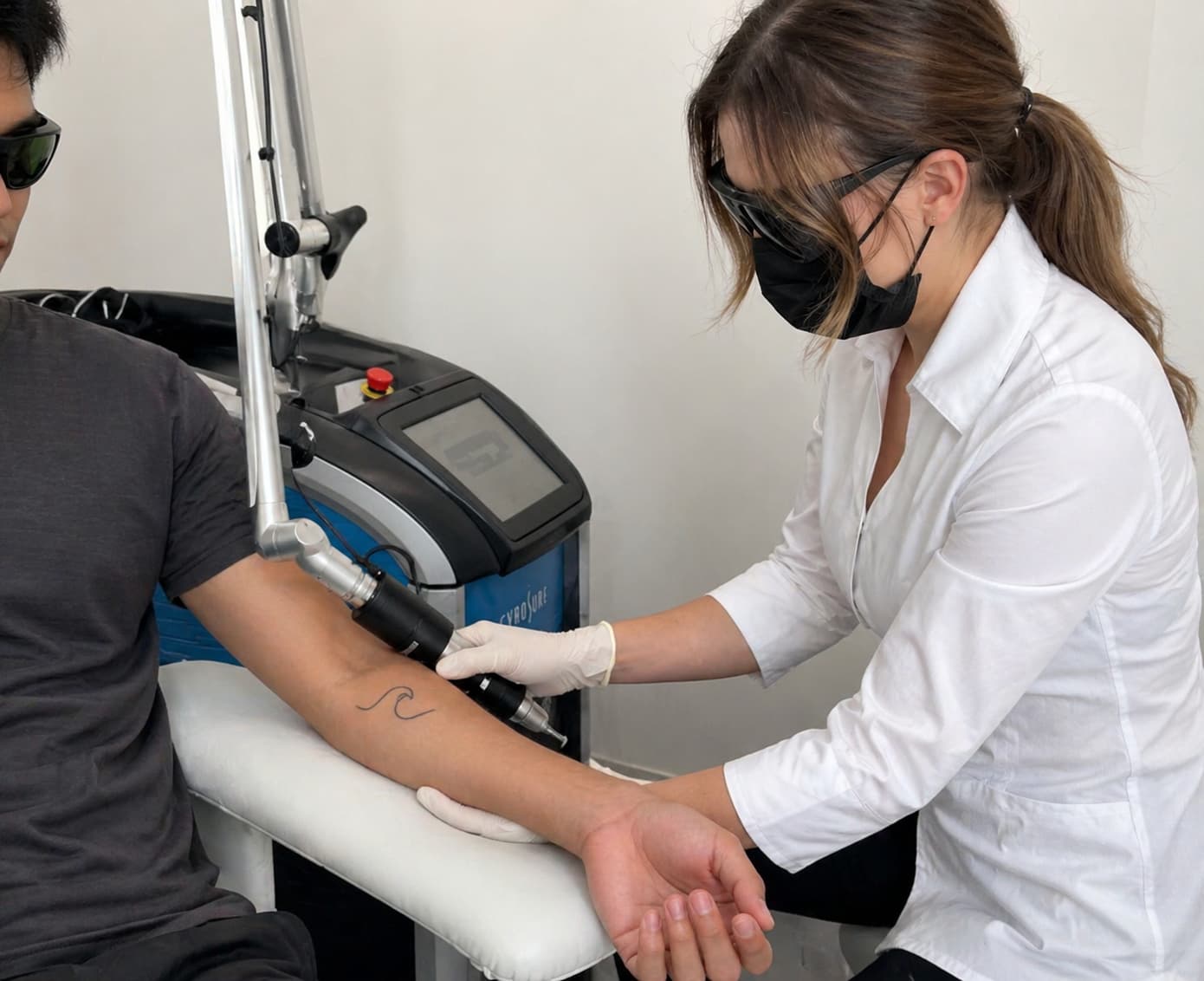 A practitioner performs laser tattoo removal Winnipeg treatment in a clean, clinical spa setting. The technician, wearing protective eyewear, a face mask, and gloves, carefully holds a handheld laser device connected to a Cynosure PicoSure machine, directing it at a client’s forearm. The client is seated with their arm resting on a padded support, wearing protective glasses while a small, simple outline-style tattoo is being treated. The room is bright and minimal, with white walls and professional equipment visible in the background, creating a focused and hygienic environment for advanced skin treatments.